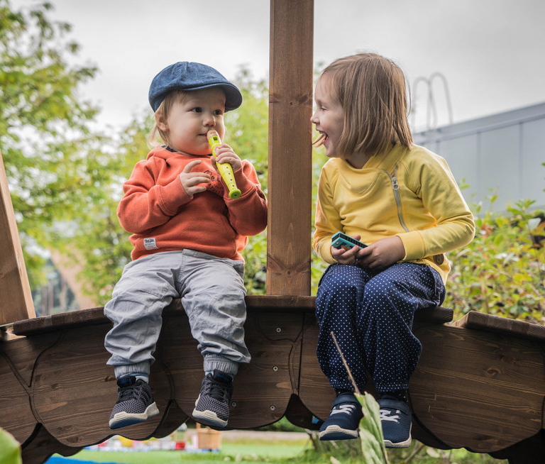 Two young children sit on a wooden bridge. The child on the left wears a blue cap, orange sweater, and gray pants, blowing a bubble wand. The child on the right wears a yellow sweater and blue pants, smiling and looking at the other child. Trees and play equipment are in the background.