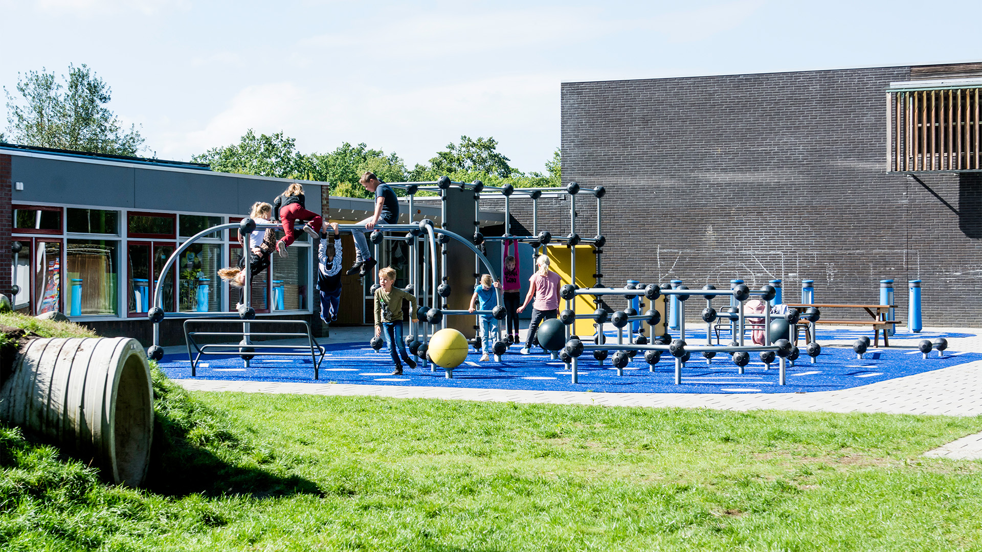 Children playing on a modern playground structure under a partly cloudy sky. The playground features various climbing and balancing apparatuses on a blue rubberized surface. The area is adjacent to a dark brick building with large windows and greenery nearby.