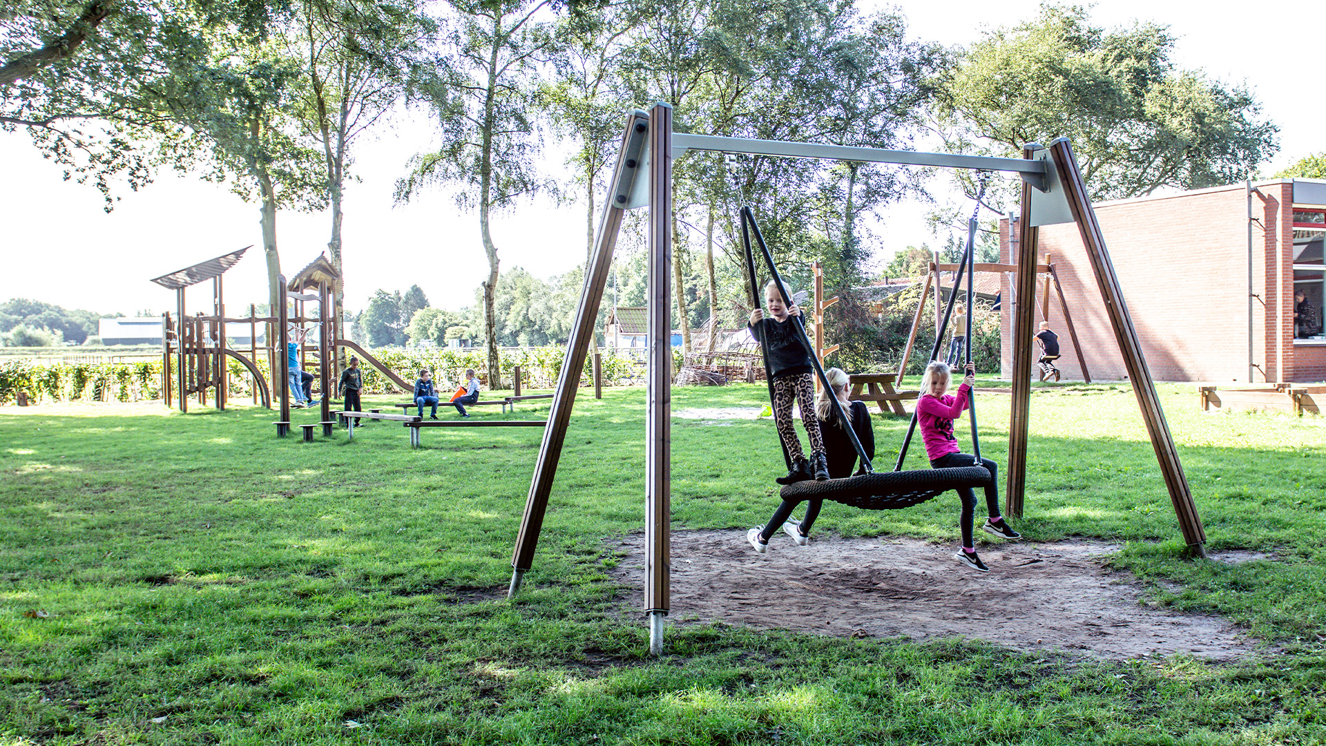 Children are playing at a playground in a park on a sunny day. There is a swing set in the foreground with two children swinging, one in black and one in pink. In the background, other kids are playing on a climbing structure and adults are nearby. Trees and grass surround the area.