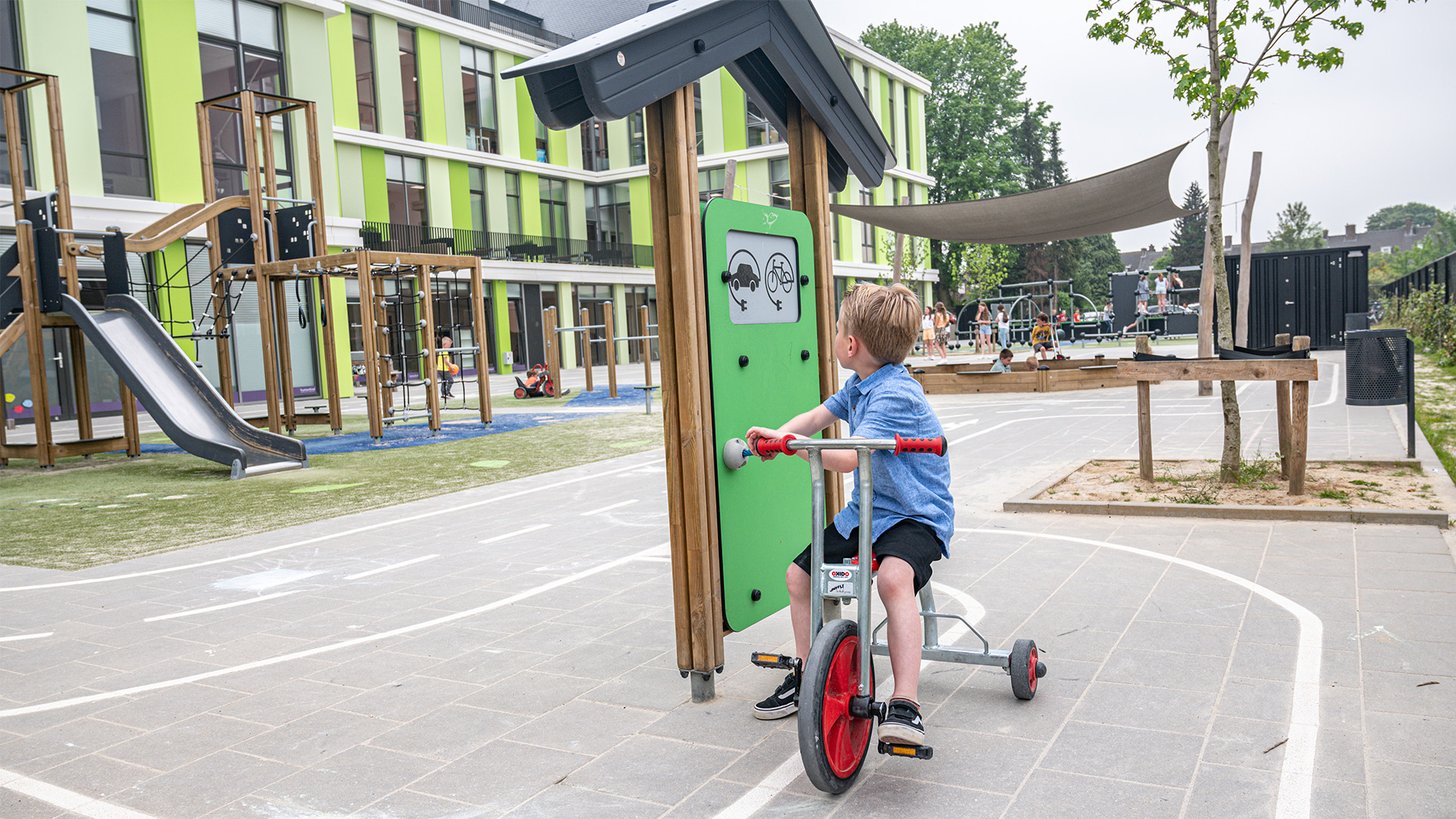 A young child rides a tricycle on the playground of a modern school. The playground has a slide, climbing structures, and a shaded area. The school building has a vibrant green and white facade with large windows. Other children are playing in the background.