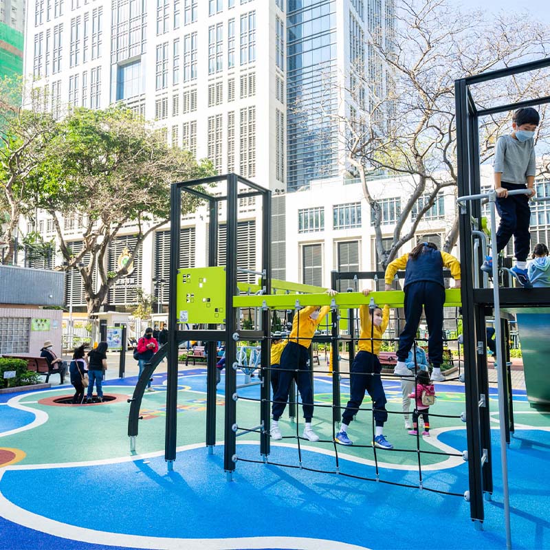 Children and adults enjoy various activities at a colorful, modern playground set against the backdrop of tall buildings. Some kids climb on equipment, while others swing or play games on the cushioned, patterned surface, under a clear sky with some trees nearby.