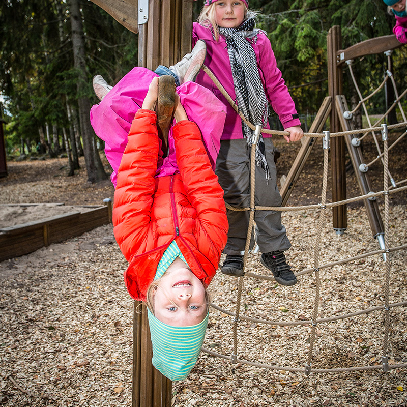 Two children are playing in an outdoor playground. One child, wearing a red jacket and green striped hat, is hanging upside down from a wooden structure, smiling. The other child, wearing a pink jacket and scarf, stands on wood chips near a rope climbing net.