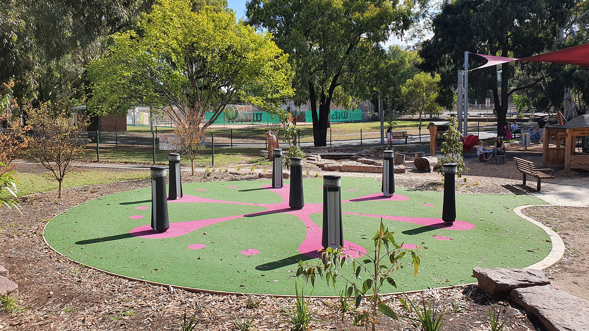 A circular play area featuring six vertical black posts on a green and pink surface. Surrounding the area are trees, a fence, and playground equipment. A few children and adults are visible in the background, enjoying the outdoor space.