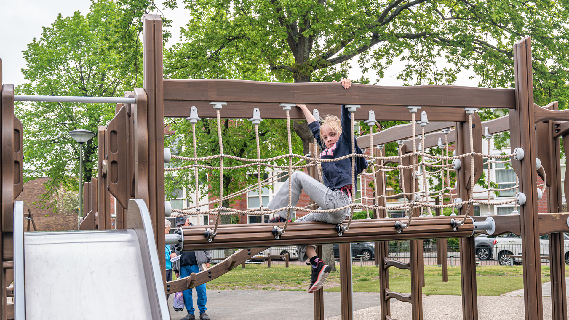 A young child with blonde hair is climbing on a rope bridge at a playground. She is holding onto the ropes with one hand while looking ahead. The playground equipment is brown, and there are trees and buildings in the background. Other children are in the distance.