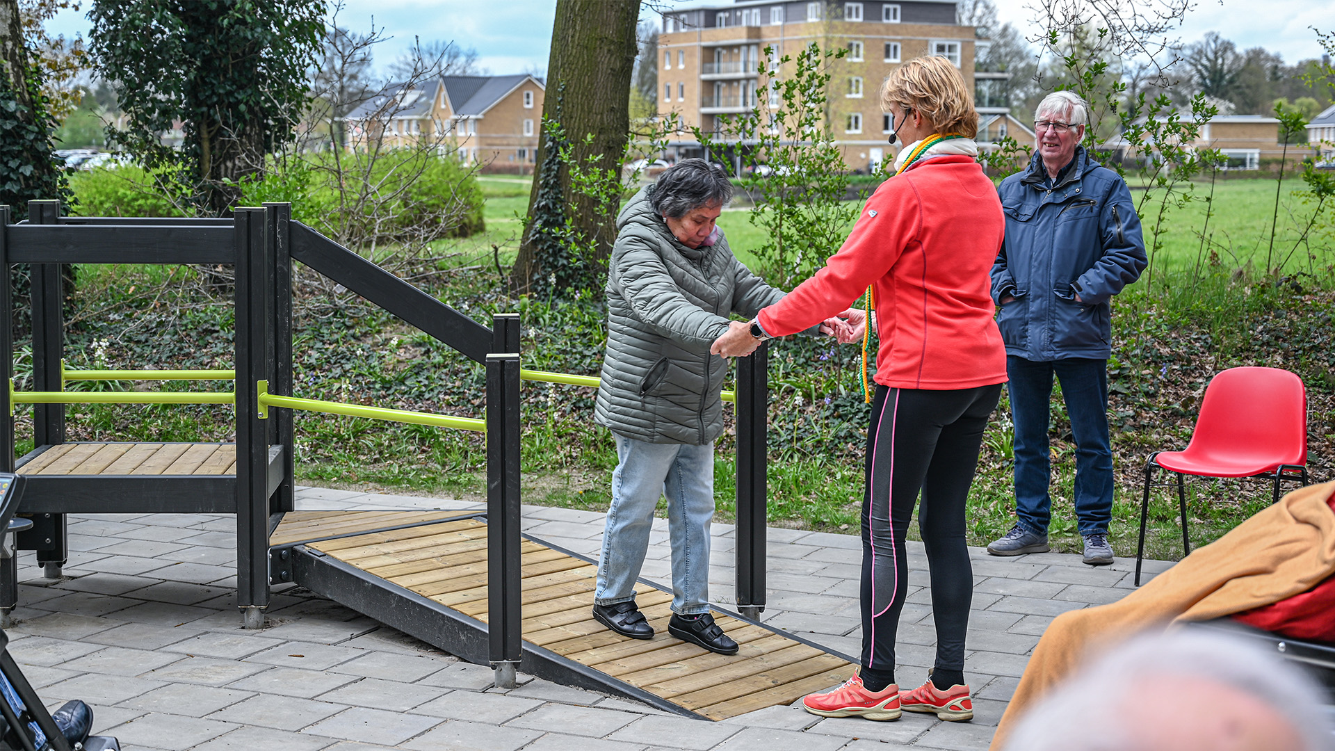 An elderly woman in a green jacket uses a wooden and metal outdoor ramp with assistance from a woman in a red jacket. A man in a blue jacket stands nearby. Trees and residential buildings are visible in the background.