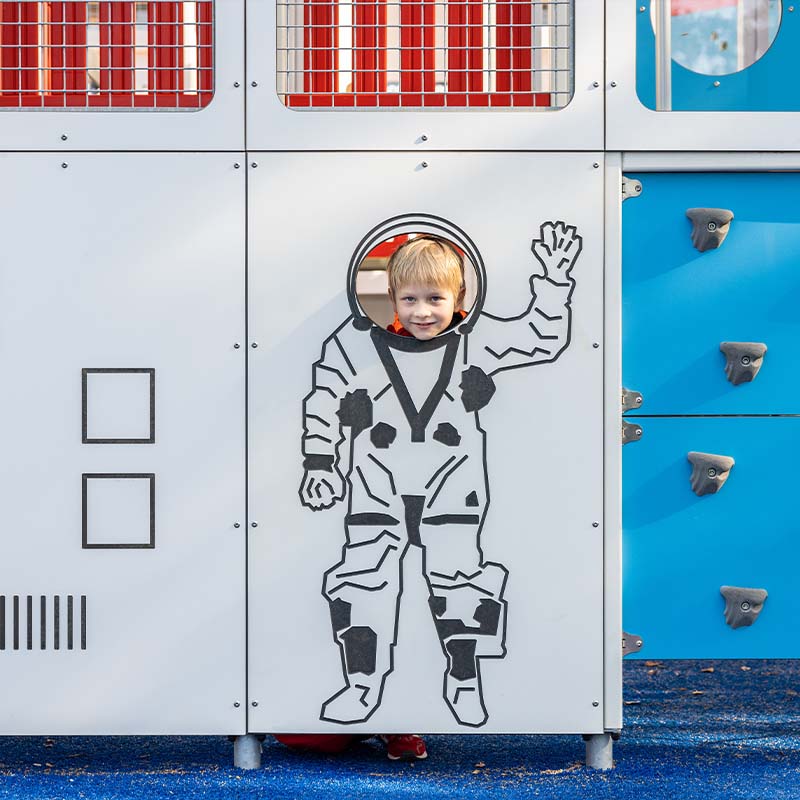 A smiling child is peeking through a cutout of an astronaut on a playground structure. The child appears to be waving, with the upper half of their body showing through the hole where the astronaut's face should be. The playground features a blue climbing wall.