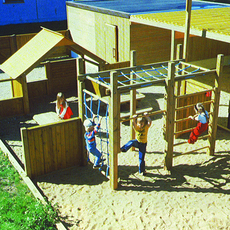 Children are playing on a wooden playground set. One child is climbing a net, another is hanging from monkey bars, and another is on a ladder. The playground is built on a sandy ground with wooden structures, including a playhouse and climbing frame.