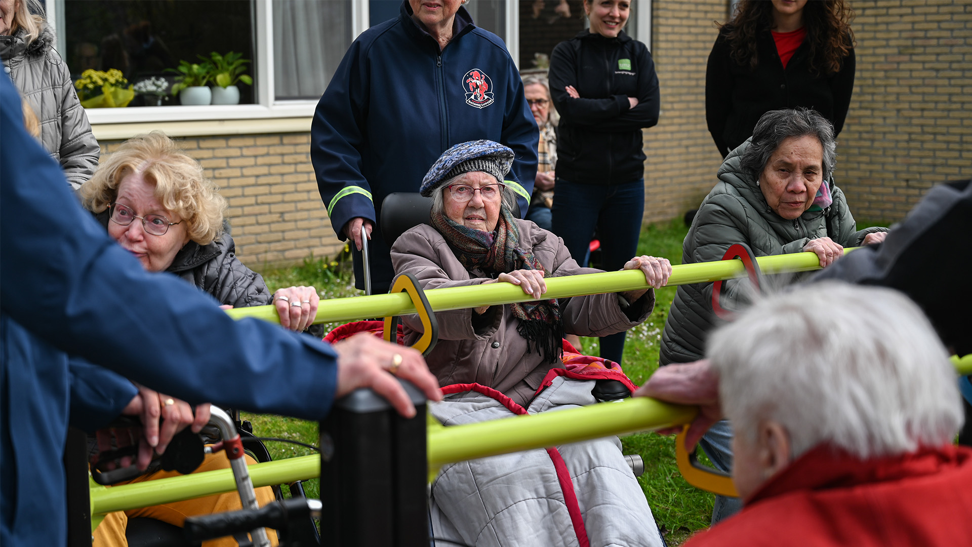 A group of elderly individuals and caregivers are gathered outside. Some are seated in wheelchairs, and two women are holding onto a yellow exercise bar. The group appears engaged and happy, with a green lawn and brick building in the background.