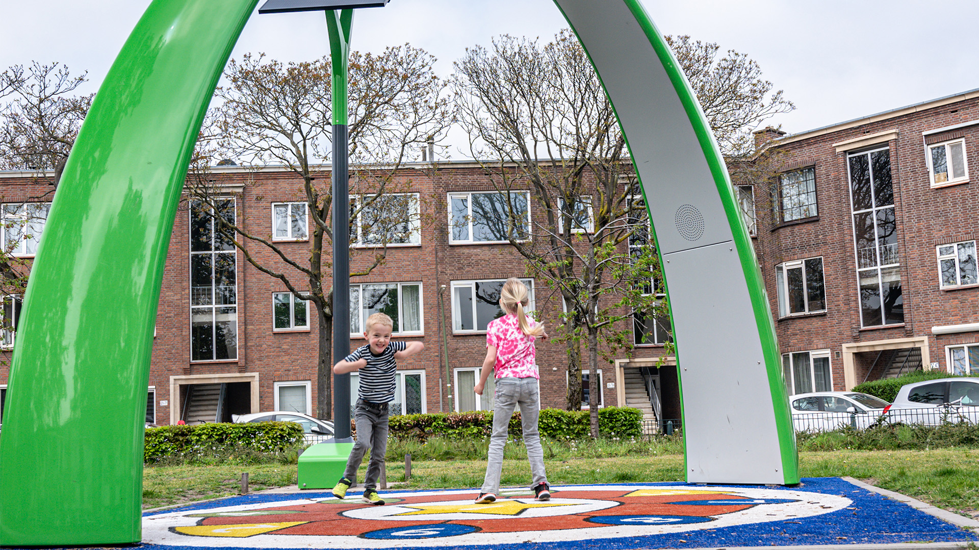 Two children are playing on a colorful, circular swing structure in a playground. The structure has green arches and is situated in a residential area with brick apartment buildings and parked cars visible in the background. Trees with sparse leaves are also in the scene.