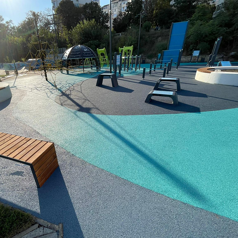 Outdoor recreational area with colorful surfaces in shades of blue and grey. Exercise equipment and playground structures, including climbing nets, a dome-shaped climbing frame, balance beams, and workout stations, are dispersed throughout the area. Trees and buildings are in the background.