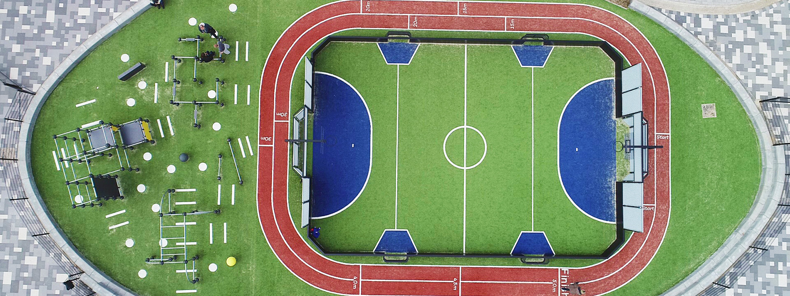 Aerial view of a sports complex featuring a green mini soccer field with blue goal areas, surrounded by a red running track. To the left, there is outdoor gym equipment, and the area is paved with grey patterned tiles.