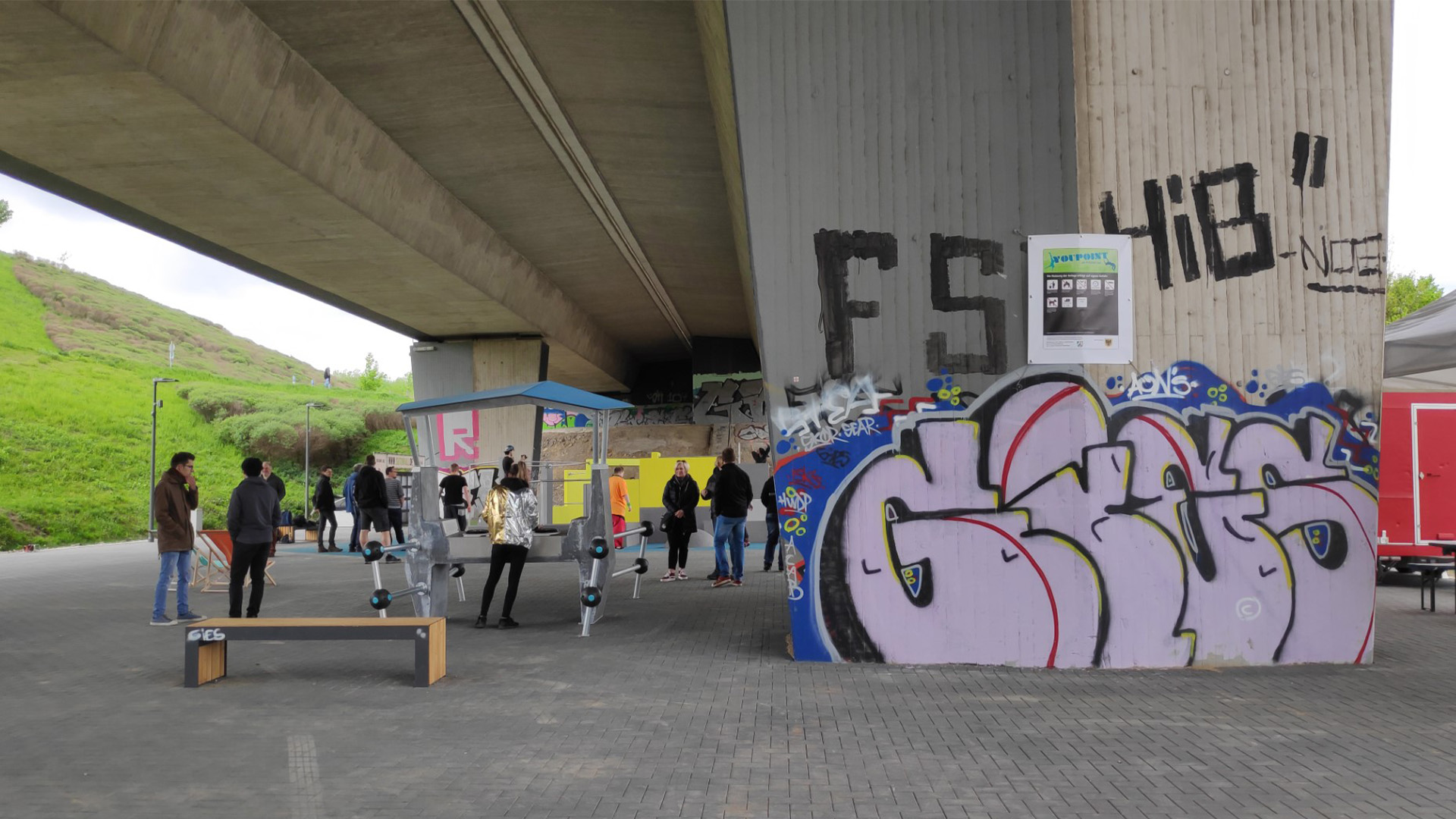People are gathered under a highway bridge near graffiti-covered pillars. Some are interacting with art installations or hanging out near a picnic bench. The setting includes a park-like area with grassy slopes in the background.