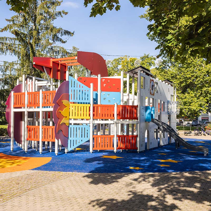 A colorful playground structure shaped like a ship, featuring multiple slides, climbing elements, and play areas. The ground is covered with blue rubber matting, and the surrounding area has trees providing shade.