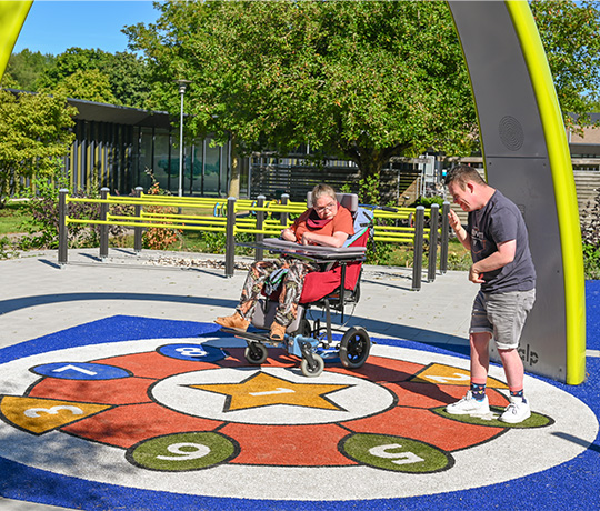 Two clients from the Ipse de Brugge healthcare facility dance under the Lappset Sona Interactive dance and play arch. An inclusive playground with multiple Lappset inclusive equipment, including the yellow Sona. 