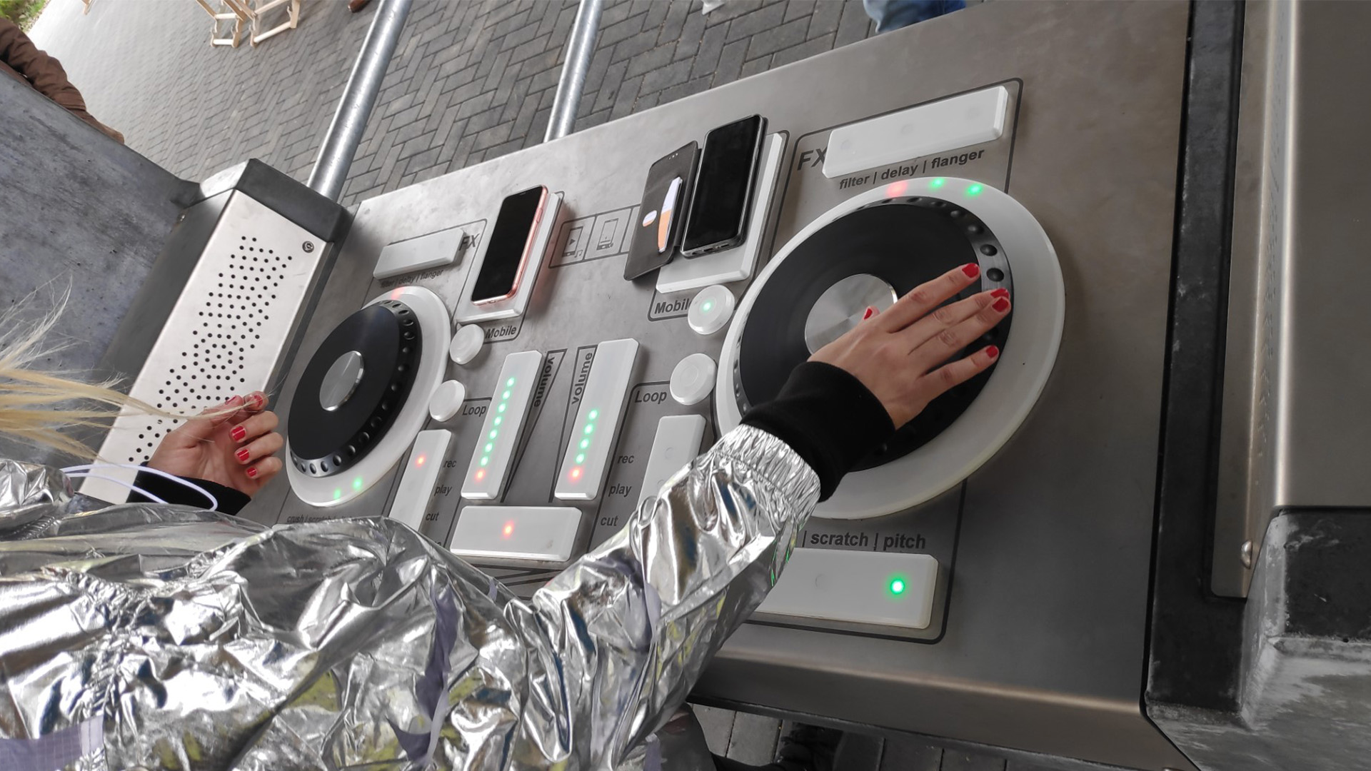 A person in a shiny silver jacket with red nail polish is using a DJ controller on an outdoor setup. One hand is placed on a turntable and the other on a set of buttons. Nearby, there are phones and equipment. The ground is paved with bricks.