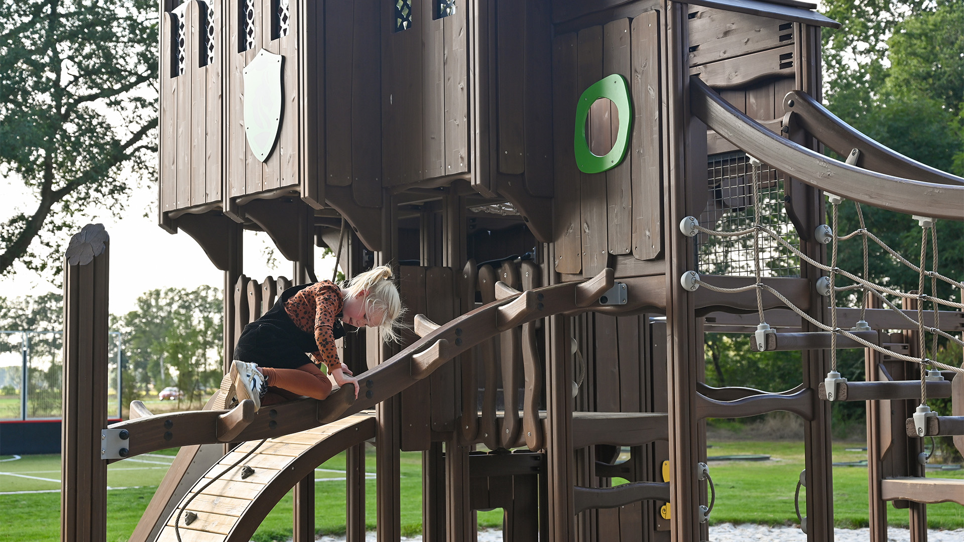 A young child with blonde hair climbs on a wooden playground structure shaped like a treehouse. The playground is set in an outdoor area with green trees in the background and white gravel beneath. The structure includes ropes and various climbing platforms.