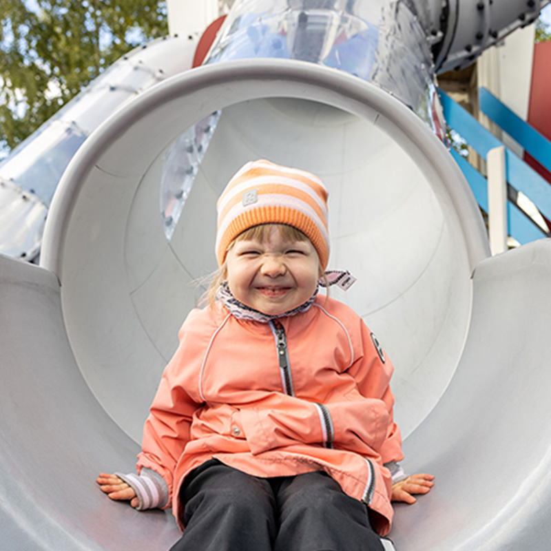 A small child wearing a pink jacket and an orange beanie is smiling and sitting at the bottom of a slide, likely at a playground. The slide appears to be a metallic, tube-like structure. Trees and part of the playground equipment are visible in the background.