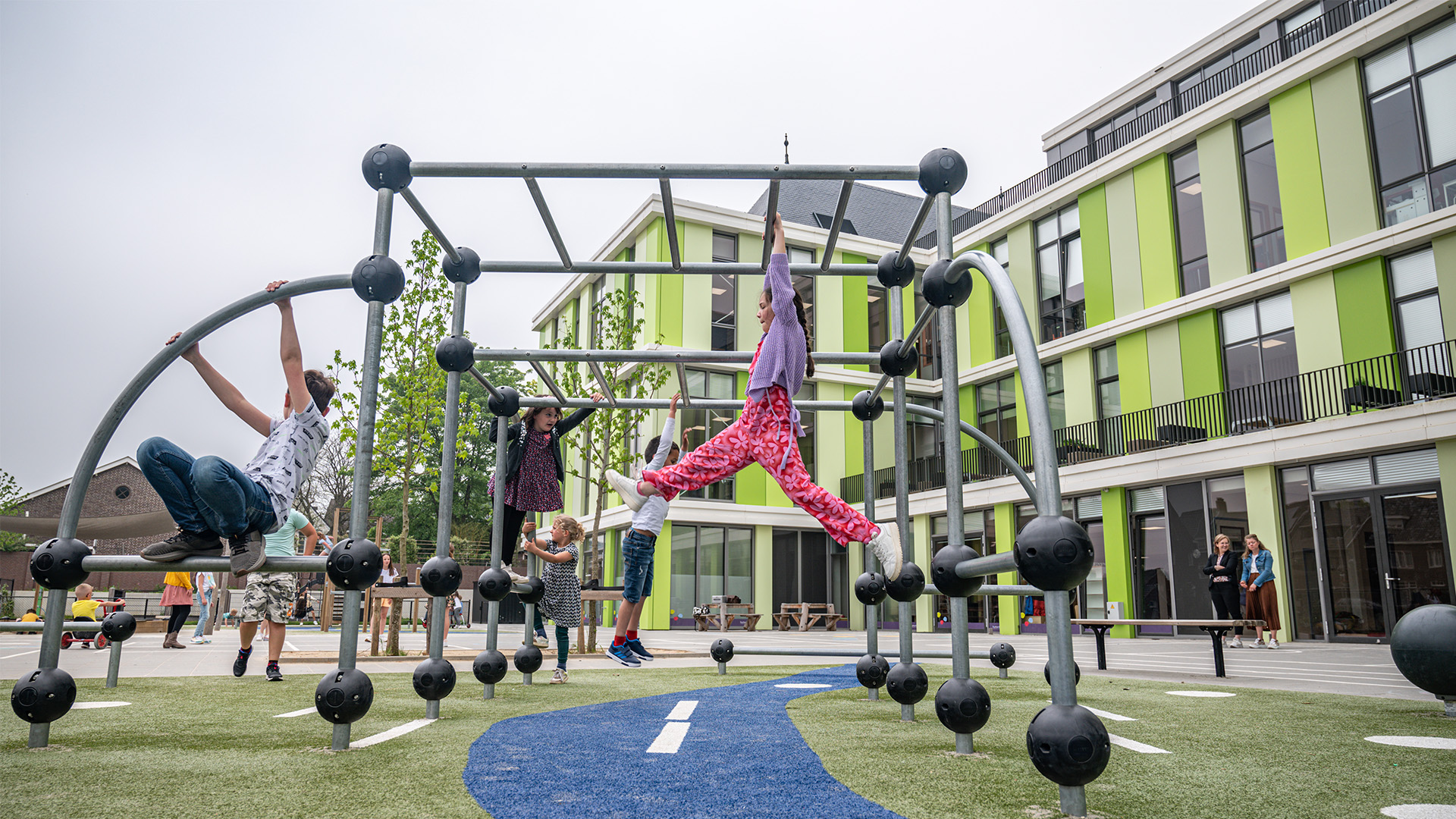 Children playfully climb on a modern jungle gym structure at a colorful outdoor playground next to a contemporary school building. Some children dangle from the equipment while others climb, enjoying an active and fun outdoor activity.