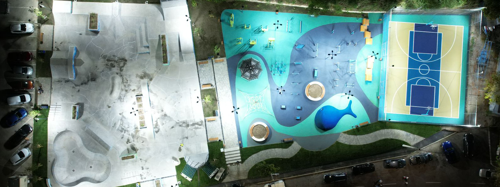 An aerial nighttime view of a brightly lit recreational area featuring a concrete skatepark on the left, a colorful playground with various equipment in the center, and a basketball court with two hoops on the right. Several cars are parked around the perimeter.