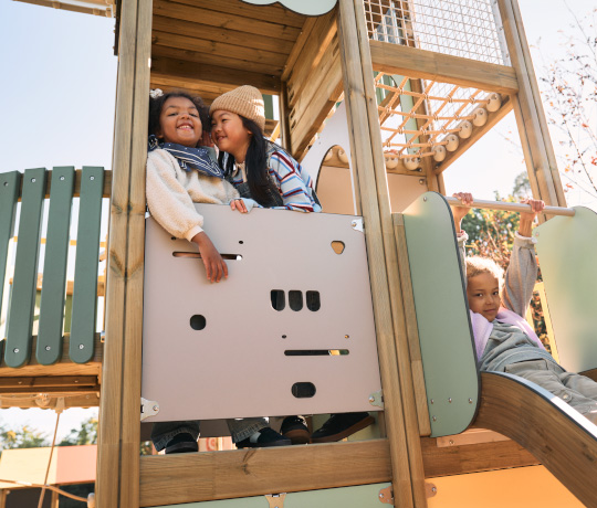 Photo from a playground where two children play in a tower and one slides down. The Lappset Prime play structure is made of wood in light tones.