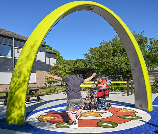 Two clients from the Ipse de Brugge healthcare facility dance under the Lappset Sona Interactive dance and play arch. An inclusive playground with multiple Lappset inclusive equipment, including the yellow Sona. 
