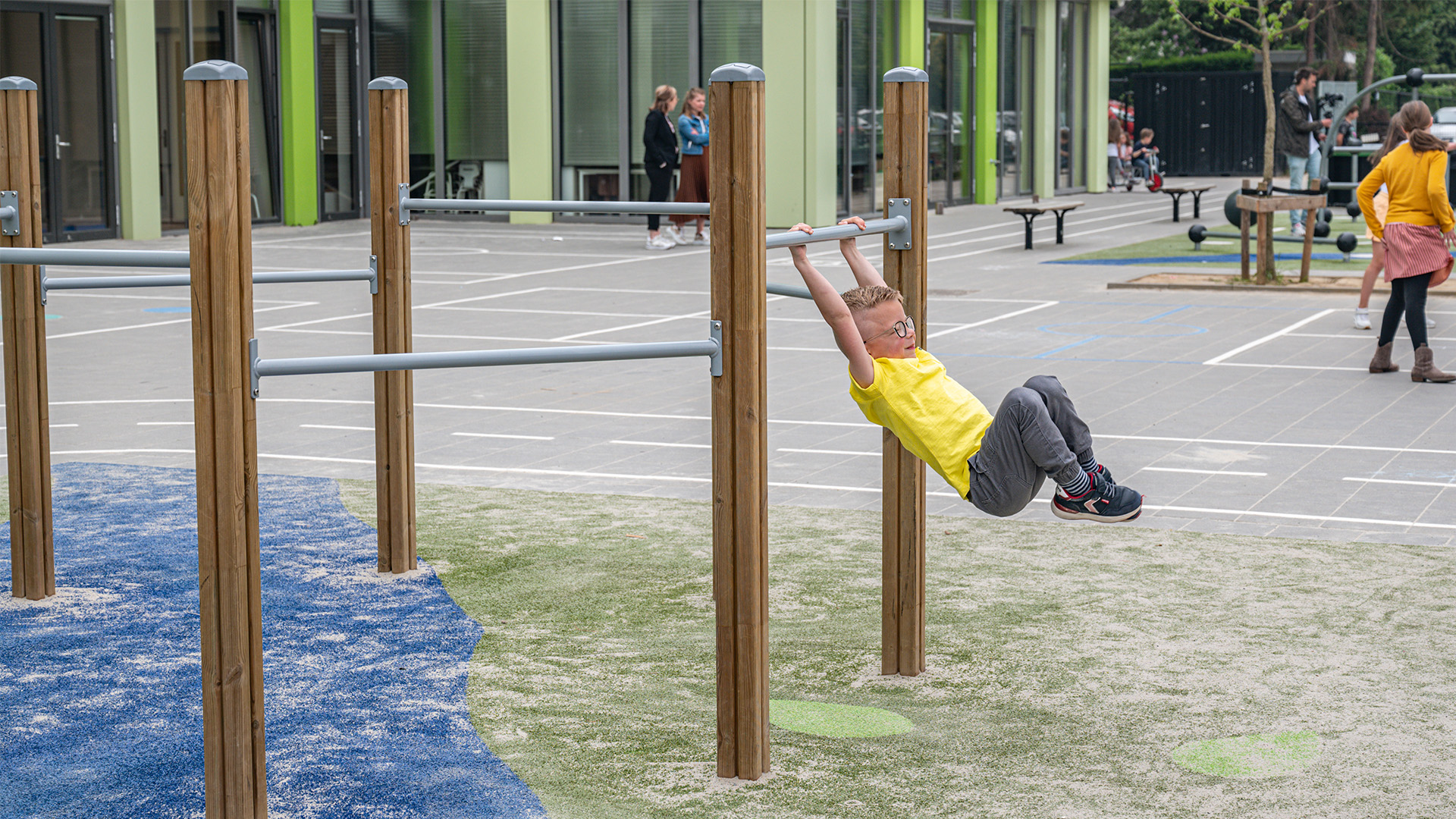 A young boy in a yellow shirt and jeans hangs by his hands from a horizontal bar at a playground. The playground has wood and metal climbing structures, colorful rubber flooring, and several other children playing in the background near a modern building.