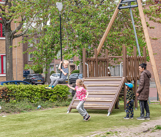 Children enjoying a day at the playground. One child is swinging on a zip line while another is running below. Two more children and an adult stand nearby, watching and waiting for their turn. The setting includes green grass, trees, and urban residential buildings.