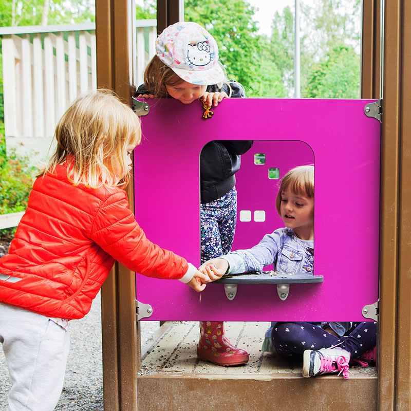 Three children are playing at a playground. Two stand on a raised platform, one in a red jacket and another wearing a cap with a Hello Kitty design. The third child, in a purple jacket and patterned pants, sits below and interacts with the others through a purple window panel.