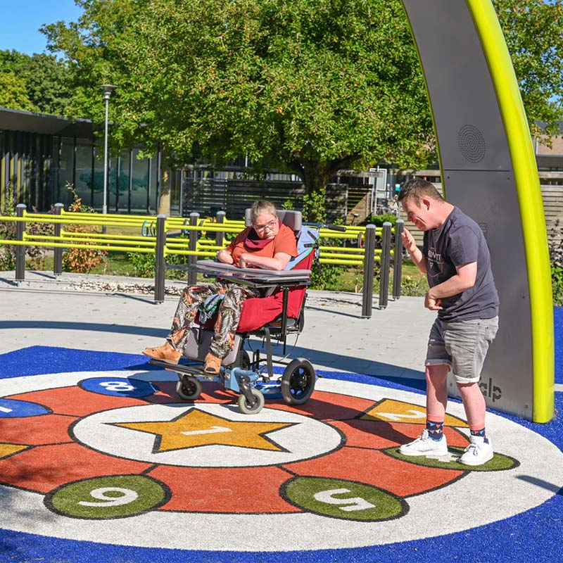 Two clients from the Ipse de Brugge healthcare facility dance under the Lappset Sona Interactive dance and play arch. An inclusive playground with multiple Lappset inclusive equipment, including the yellow Sona. 