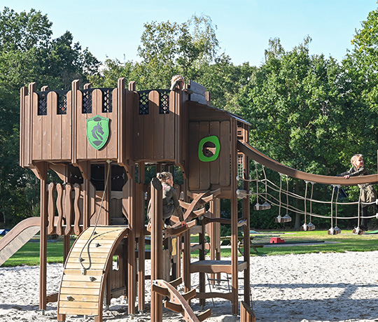 A wooden playground structure resembling a castle with various climbing elements, including rope bridges, ladders, and slides. The playground is set in an outdoor park with trees and grass in the background. Children are seen playing on the structure.