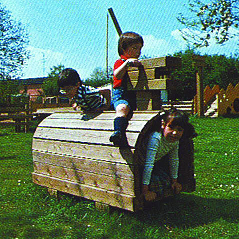 Three children play on a wooden structure shaped like a train engine in a grassy area with trees and a playground in the background. One child is climbing, another is sitting on top, and the third is crouching inside.