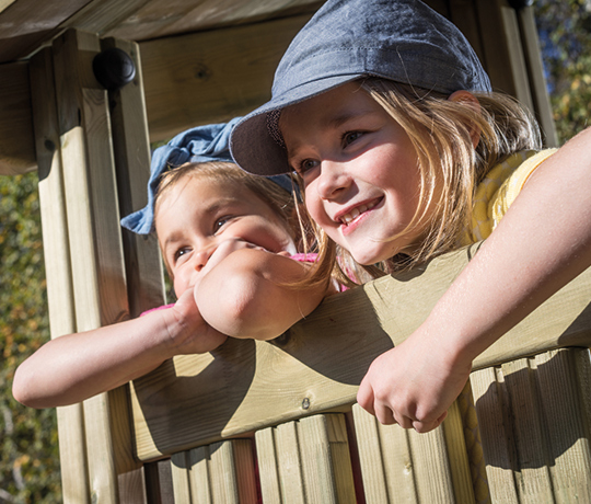 Two smiling children lean over a wooden railing outside, one wearing a light blue cap and the other a blue headband. They appear happy and relaxed, enjoying a sunny day of nature play. 