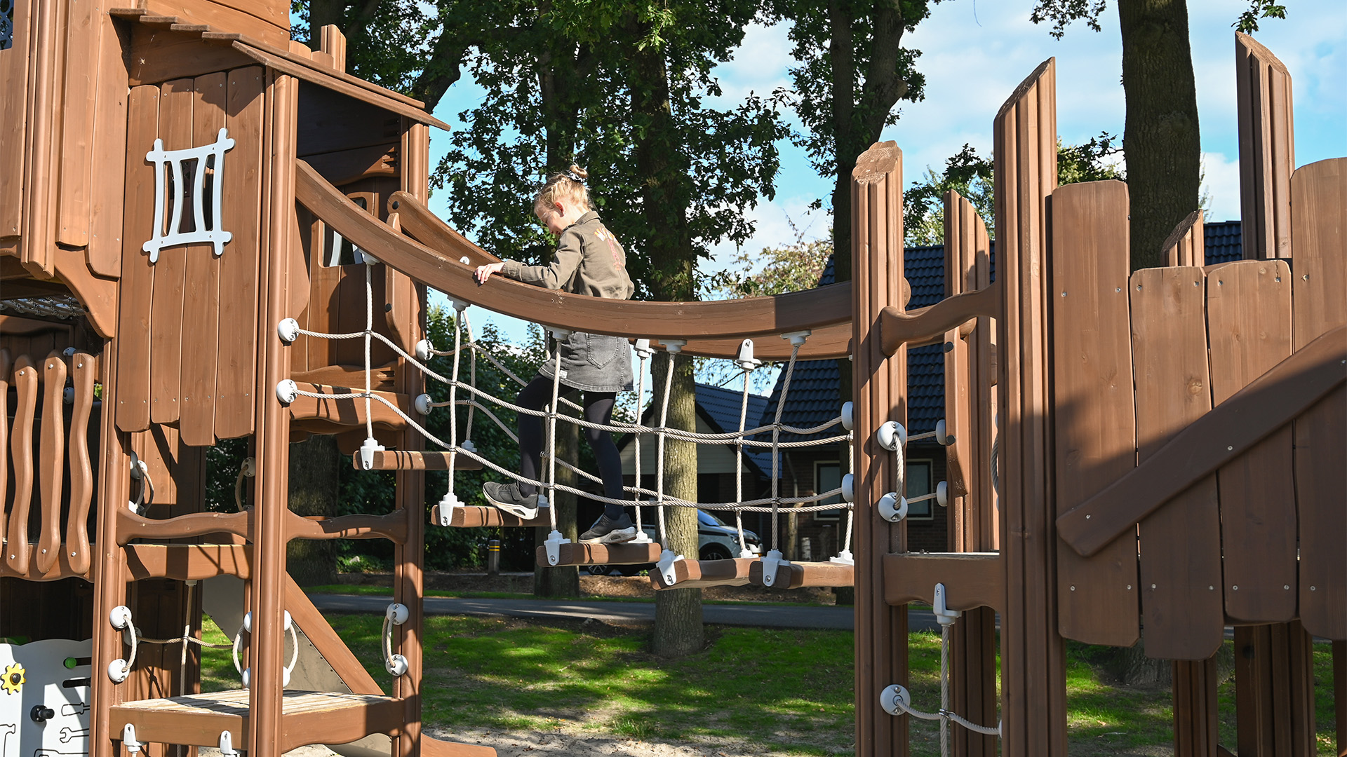 A child in a green jacket and black pants is walking across a wooden rope bridge in an outdoor playground. The playground structure is made of wood and features various climbing and balancing elements. Trees and a house are visible in the background.