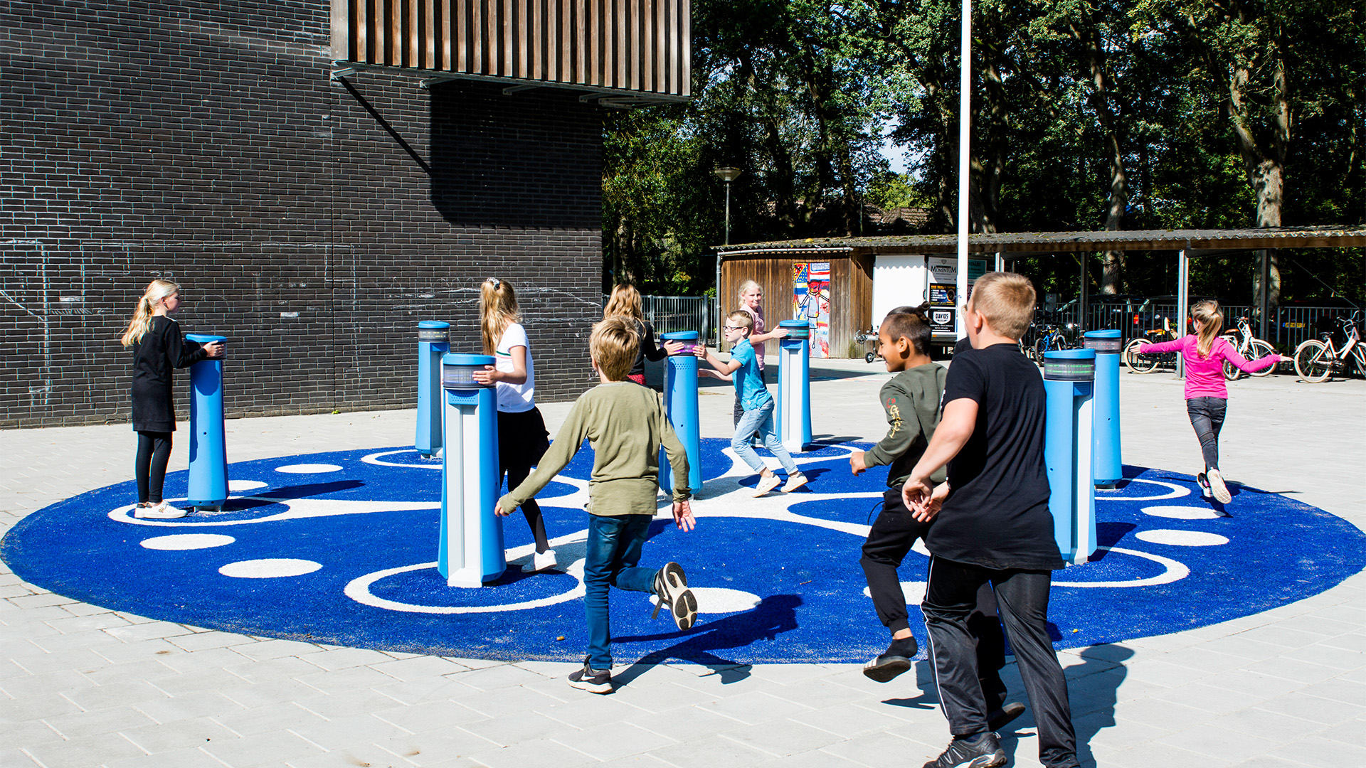 Children are playing on a colorful interactive playground. The playground features blue pillars on a circular blue mat with white spots. A brick building and trees are in the background. The children appear to be enjoying their time, running and interacting with the equipment.