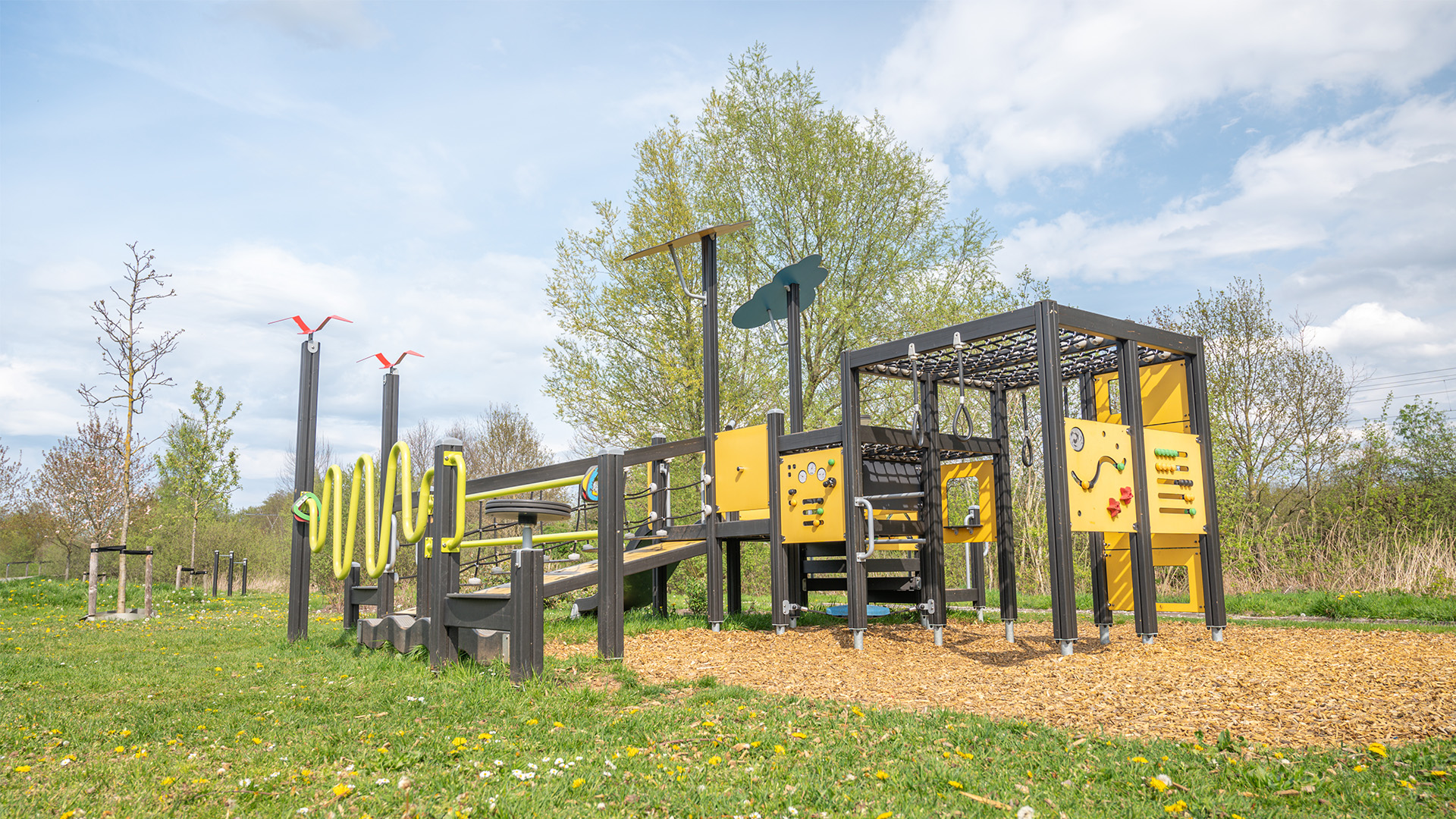 An outdoor playground with various climbing structures, slides, and activity panels, all on a woodchip-covered ground. The play equipment is set in a grassy area with trees and a clear sky in the background.
