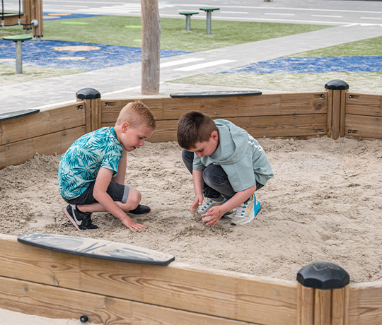 Two young boys are playing together in a sandbox at a playground. One boy is wearing a green leafy-patterned shirt, while the other is wearing a plain green shirt. They are both focused on building something in the sand. The playground is in the background.