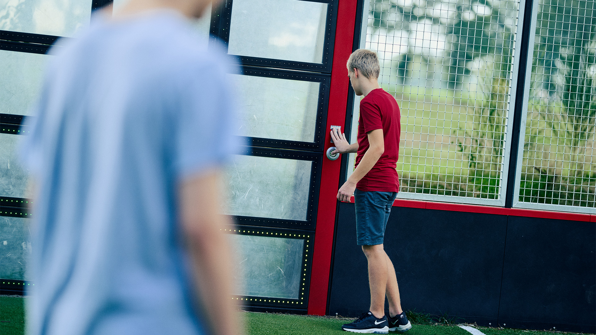 A young person wearing a red shirt and gray shorts is opening a door with a metal handle. Another person in a blue shirt is walking away in the foreground, slightly out of focus. The background features wire mesh panels and greenery.