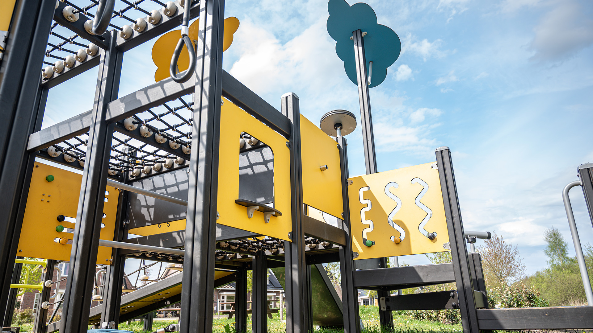 A colorful playground structure features various climbing nets, a slide, and interactive panels with shapes and symbols. The equipment is mainly yellow and black, set against a backdrop of trees and a blue sky with clouds.