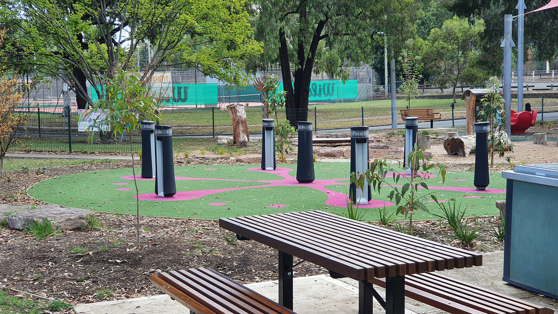 Outdoor recreational area with a wooden picnic table in the foreground and a play area featuring multiple vertical poles on a green and pink rubber surface in the background. Trees and plants are scattered around, with a tennis court visible in the distance.