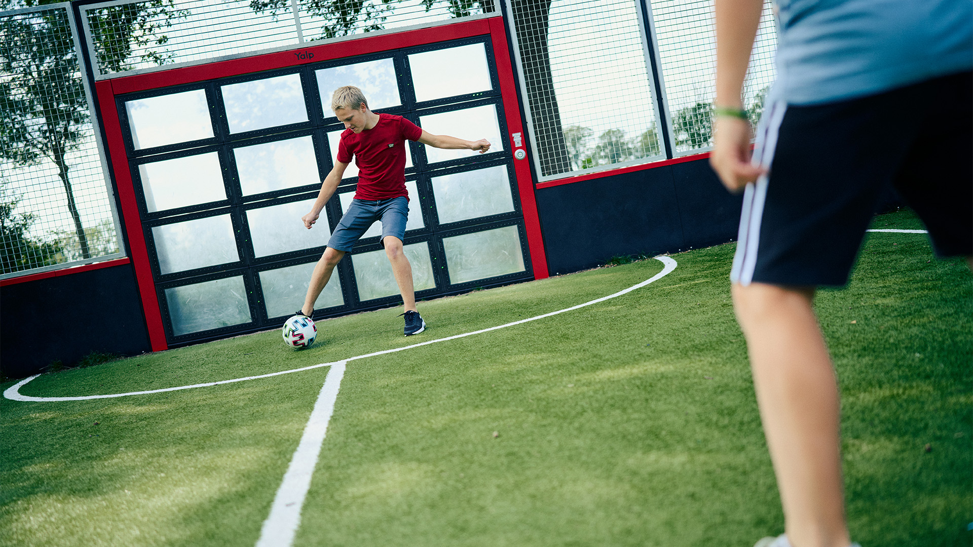 Two people are playing soccer on a small outdoor field. One person in a red shirt is kicking the ball towards the goal, while the other person in a light blue shirt stands facing them, ready to defend. The goal is framed with red and black colors.