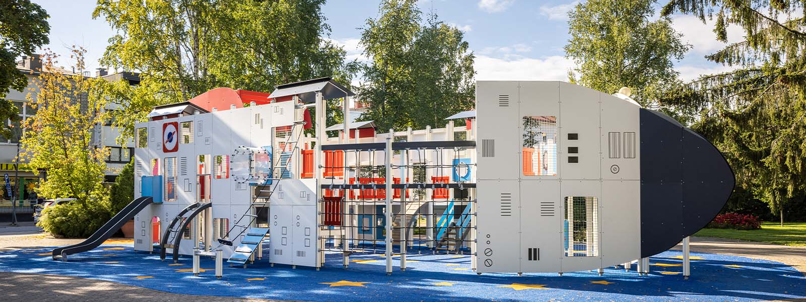 A playground structured to resemble a space shuttle and a control tower. The equipment includes slides, climbing bars, and various panels for interactive play. The ground is decorated with stars, and trees and buildings are visible in the background.