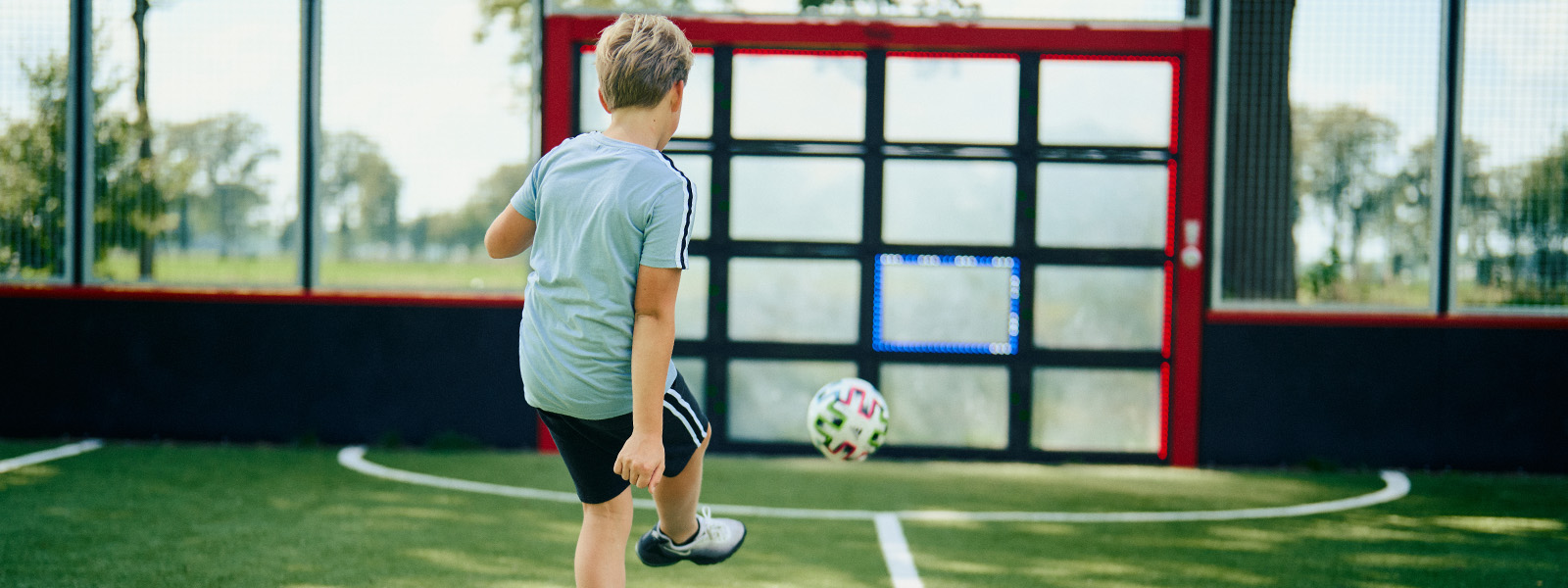 Young child playing soccer, kicking a ball towards Lappset Sutu interactive ball wall