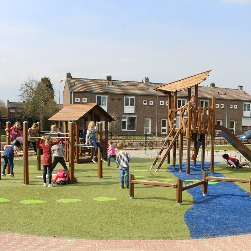 Children are playing on a playground with climbing structures and slides. The playground features a grassy area with some blue surfaces, and there are several wooden play structures. Houses and a clear, blue sky are visible in the background.