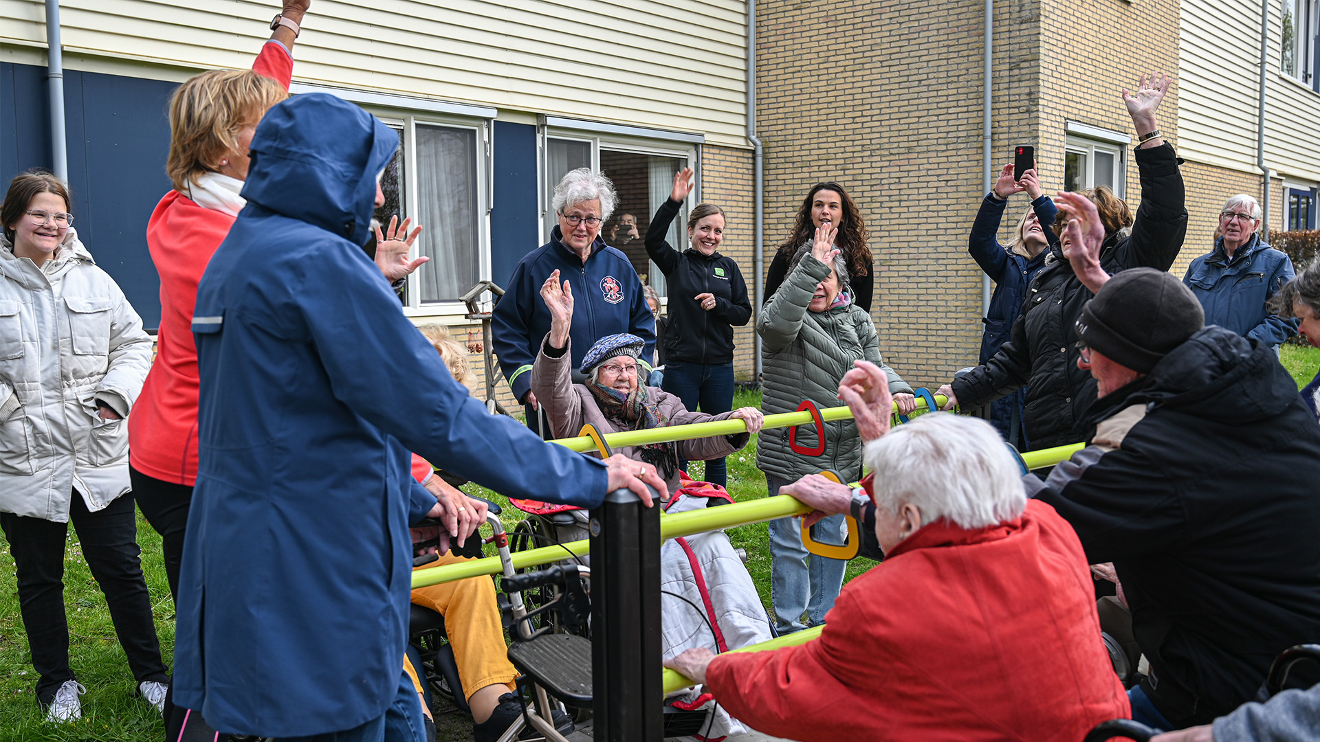 A group of elderly individuals and caregivers gather outdoors, some in wheelchairs, laughing and raising their hands. They are in front of a residential building, with grass underfoot. The mood is cheerful, and people are dressed warmly for cool weather.