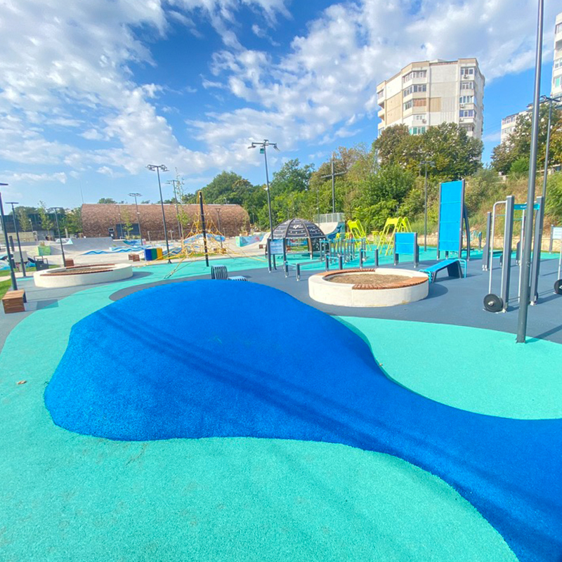 A vibrant, brightly colored playground featuring blue and green padded surfaces, various climbing structures, swings, and slides. Tall apartment buildings are visible in the background, with a clear blue sky and scattered white clouds overhead.