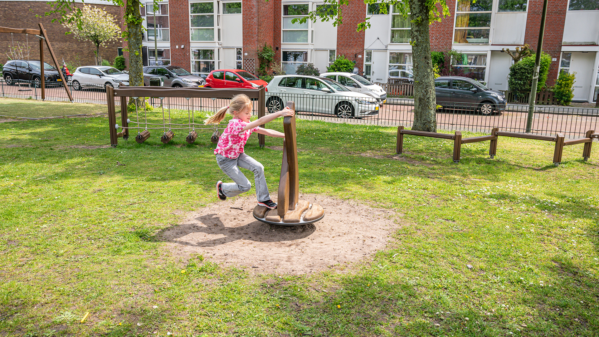 A girl in a red and white shirt and gray pants is playing on a circular spinner in a playground. The playground is grassy, and there are several pieces of equipment, including swings and balance beams. In the background, there is a row of parked cars and buildings.