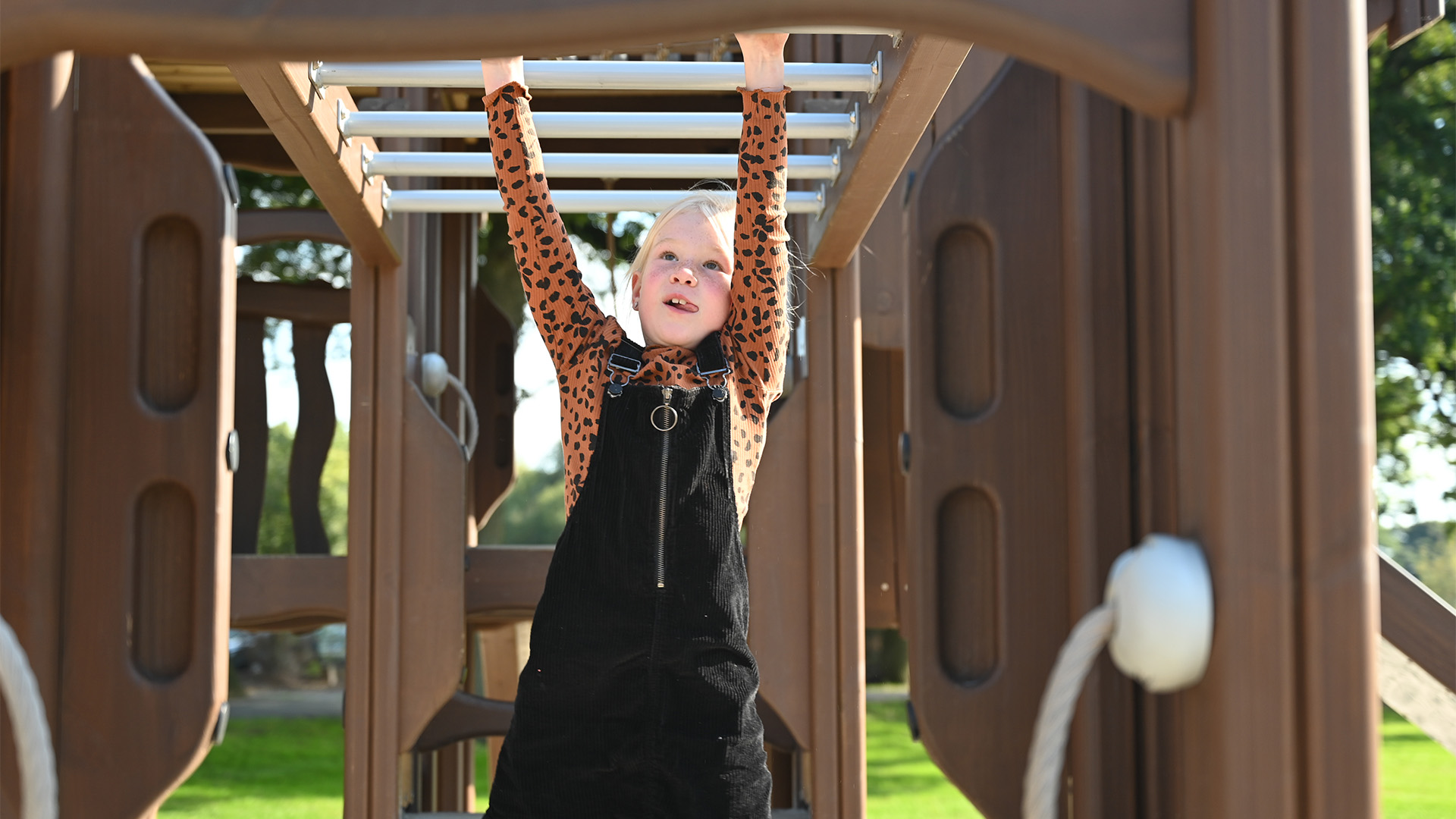 A young child with light hair in a black overall dress and long-sleeve orange polka dot shirt plays on monkey bars at a playground. They are smiling and looking up as they hang from the bars with both hands. The background is bright with green grass and trees.