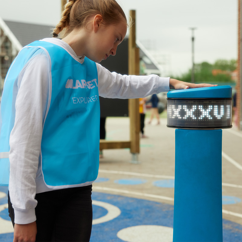 Child interacting with the Lappset Memo during Outdoor Classroom activities, wearing a blue 'Lappset Explorer' vest.