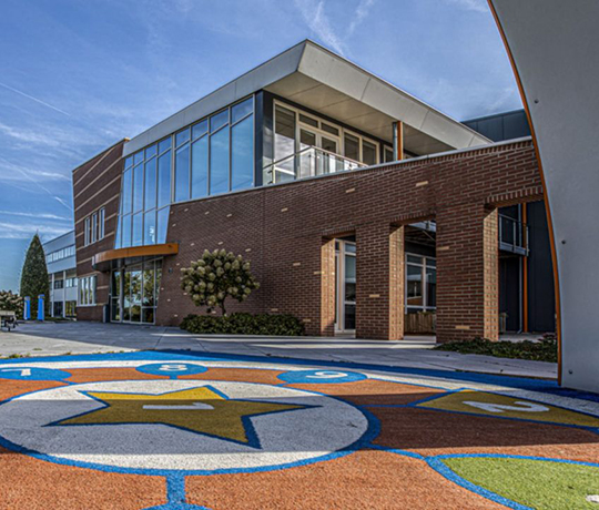 A modern brick and glass building is shown with large windows. The foreground has a colorful outdoor play area featuring a large star surrounded by numbered circles. The sky is clear and blue, indicating a sunny day.