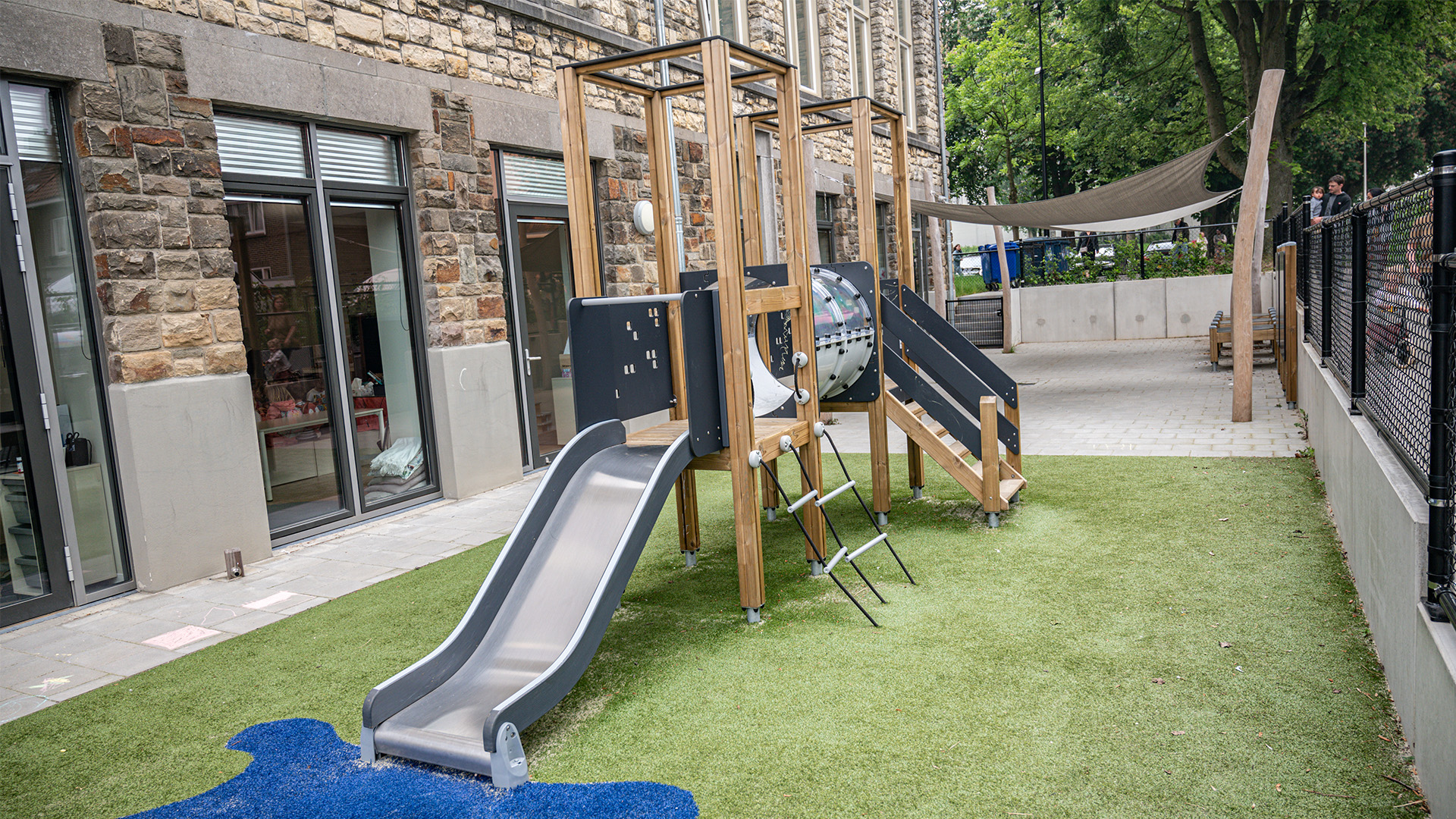 A modern playground with a metal slide, climbing ladder, and play structure featuring tunnels and platforms. The area has artificial grass, a blue play surface, and is located next to a building with large windows. Trees and a shaded area are visible in the background.